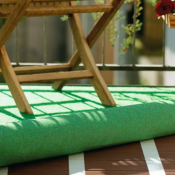 Tesa Cinta de doble cara Extra Fuerte Moquetas - 50 mm x 10 m, Blanco - Rollo de césped artificial verde en un balcón, parcialmente desenrollado debajo de una mesa de madera.