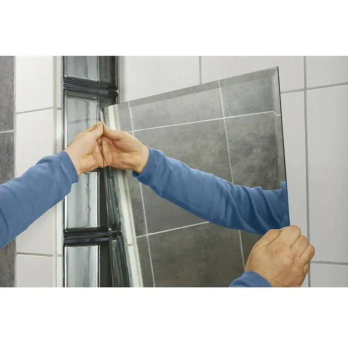 Tesa Powerbond Cinta de doble cara Espejos - 19 mm x 5 m, Blanco - La imagen muestra un espejo rectangular con bordes biselados que se está montando en una pared con azulejos grises.