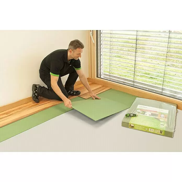 Steico Holzfaserplatte ecosilent - 790 x 590 mm, Stärke: 7 mm, 15 Stk. - Ein Mann verlegt grüne STEICO Unterbodenplatten aus Naturholzfaser unter Holzboden. Eine verpackte Packung ist ebenfalls sichtbar.