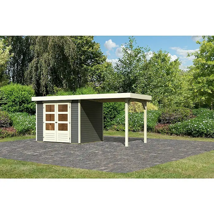 Karibu Gartenhaus Cuno 2 - Außenmaß inkl. Dachüberstand (B x T): 491 x 238 cm, Holz, Terragrau, Mit Seitendach - Das Foto zeigt ein Gartenhaus mit einem Carport-Anbau auf einer gepflasterten Fläche, umgeben von Bäumen und Sträuchern. Das Haus ist grau mit weißen Türen.
