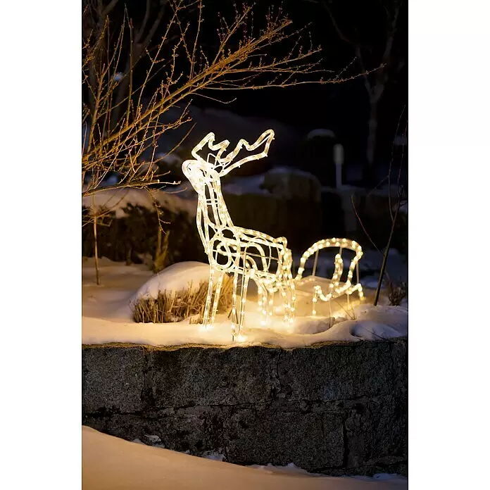 Tween Light LED-Lichtschlauch Rentier mit Schlitten - Anwendungsbereich: Außen, Warmweiß - Beleuchtete weiße Rentier- und Schlitten-Weihnachtsdekoration, warmgelb leuchtend auf einer schneebedeckten Steinmauer bei Nacht.