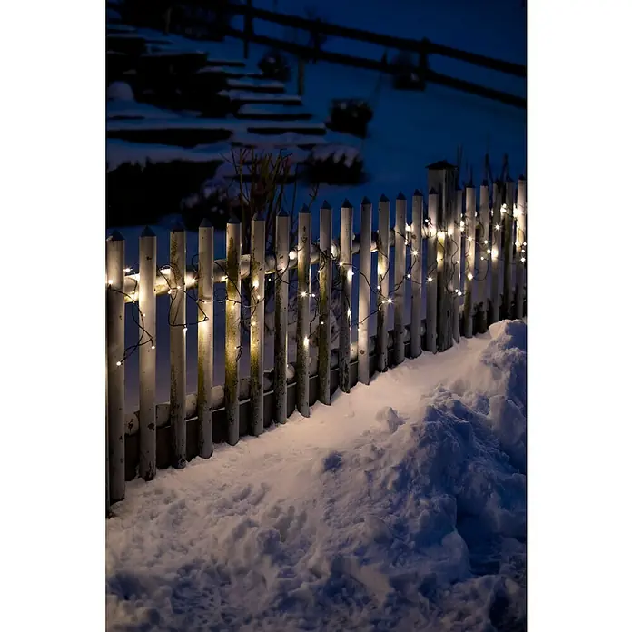 Tween Light Guirnalda luminosa LED - Para exterior, Color de luz: Blanco cálido, Número de bombillas: 120 ud., Funcionamiento en red, 23 m - Una valla blanca está adornada con luces de hadas blancas cálidas y rodeada por una gruesa capa de nieve, creando un ambiente invernal.