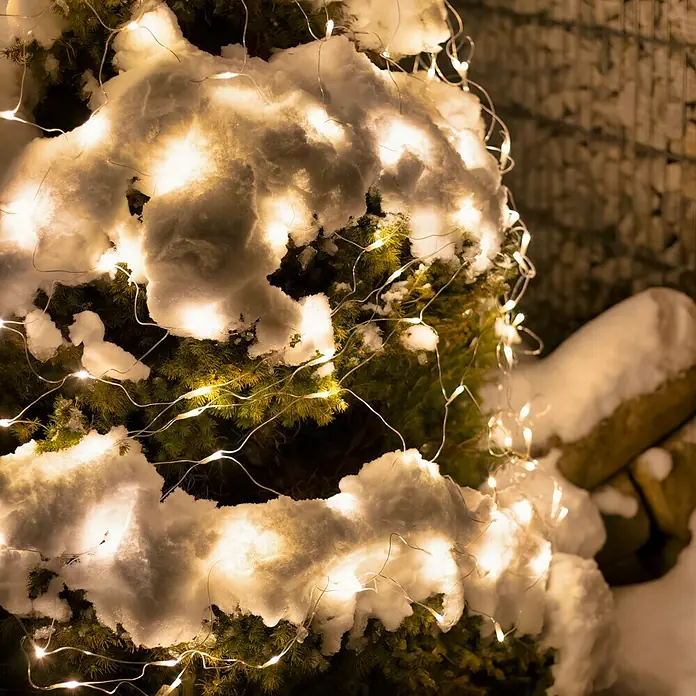 Tween Light LED-Lichtnetz - Außen, 2 m, Lichtfarbe: Warmweiß, Netzbetrieben - Nahaufnahme eines mit Schnee bedeckten immergrünen Baumes, dekoriert mit warmweißen Lichterketten, die eine gemütliche Winteratmosphäre erzeugen.