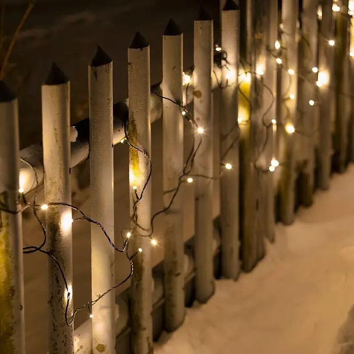 Guirnalda luminosa LED - Para exterior, 15 m, Color de luz: Blanco cálido, Número de bombillas: 192 ud., Funciona con pilas - Luces de cadena decorativas adornan una valla blanca en la nieve, creando un ambiente festivo en el espacio exterior.