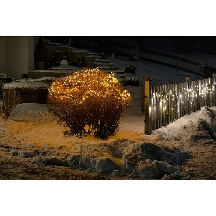 Tween Light Cadena de luces LED - Para exterior, Largo: 19,8 m, 2.000 ud., Funcionamiento en red, Color de luz: Blanco cálido - Escena de jardín invernal con un arbusto iluminado y una valla adornada con luces, creando un ambiente festivo.