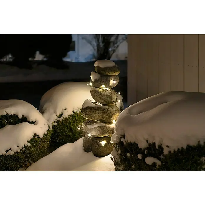 Guirnalda luminosa LED - Para exterior, 15 m, Color de luz: Blanco cálido, Número de bombillas: 192 ud., Funciona con pilas - Escena invernal festiva con una torre de piedras apiladas, adornada con luces cálidas y cubierta de nieve, creando un ambiente exterior sereno.
