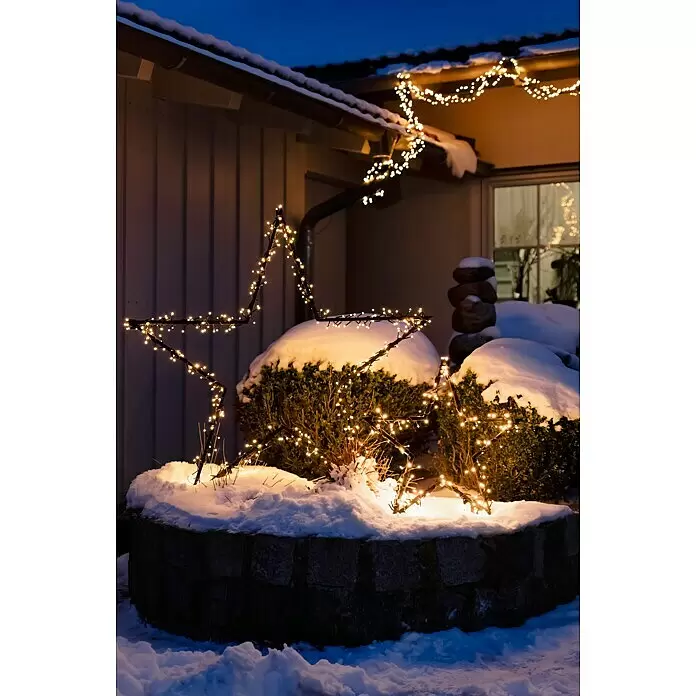 LED-Gartenstecker Stern M - Außen, Netzbetrieben, Höhe: 102 cm, Warmweiß - Eine nächtliche Außenszene zeigt eine große, warmweiße beleuchtete Sternendekoration in schneebedeckten Büschen, mit Lichterketten an der Dachrinne.