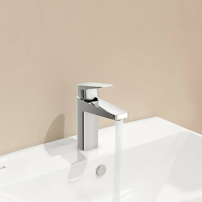 VitrA Waschtischarmatur Root Square - Chrom - Nahaufnahme eines modernen, verchromten Wasserhahns mit fließendem Wasser auf einem weißen Waschbecken vor einem beigen Hintergrund.