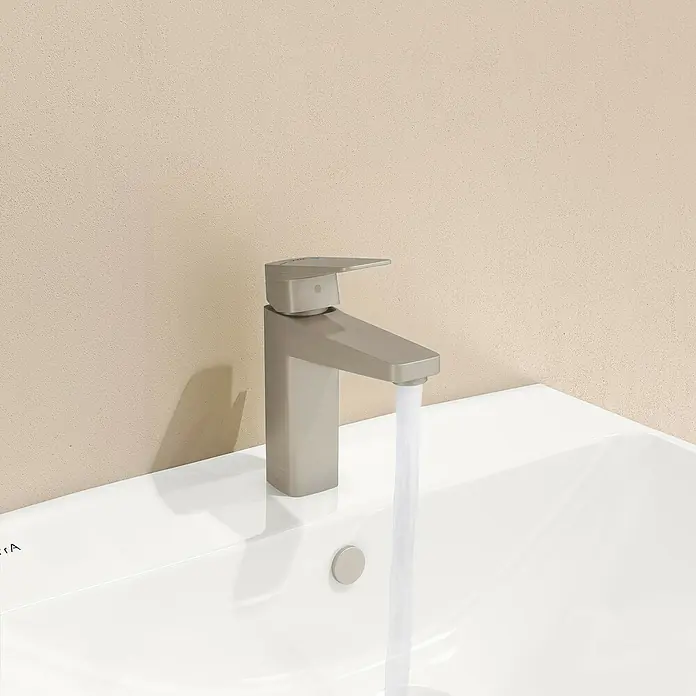 VitrA Waschtischarmatur Root Square - Nickel - Ein moderner, silberner Wasserhahn auf einem weißen Waschbecken mit fließendem Wasser vor einem beigen Hintergrund.