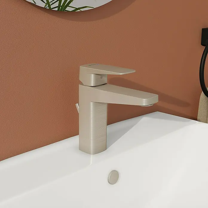 VitrA Waschtischarmatur Root Square - Nickel - Nahaufnahme eines modernen, gebürsteten Nickel-Wasserhahns auf einem weißen Waschbecken vor einer terrakottafarbenen Wand.