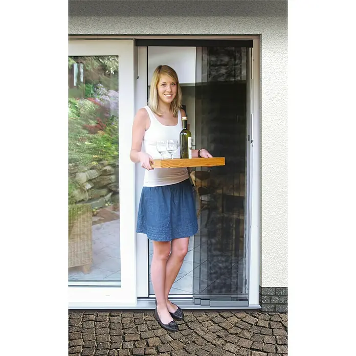Schellenberg Insect Stop Mosquitera Premium - An x Al: 95 x 220 cm, Pegado, Color bastidor: Antracita - Mujer con una bandeja de vino y copas, pasando por una puerta mosquitera. Esta puerta ofrece protección contra insectos y aire fresco.