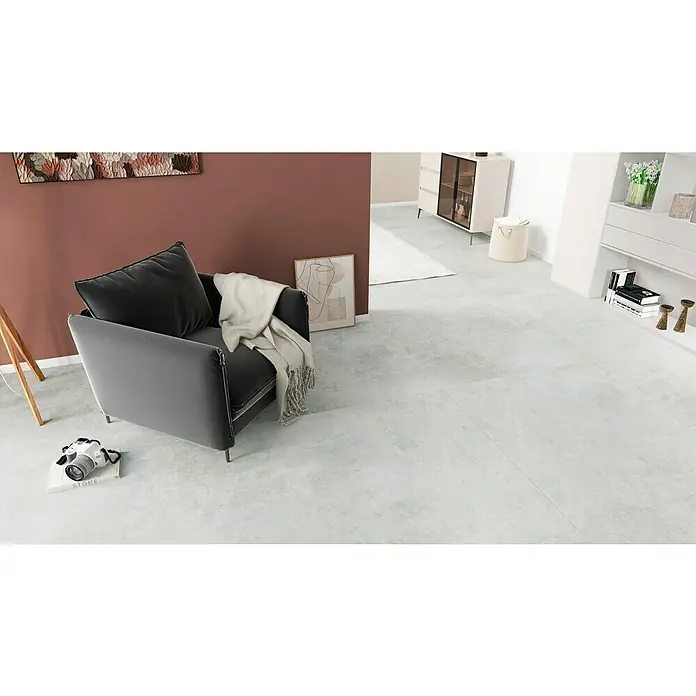 Feinsteinzeugfliese Luna Bianco120 x 240 cm, Weiß/Grau, Glänzend - Ein grauer Sessel mit Kissen und Decke, eine Kamera auf einem Buch im modernen Wohnzimmer mit Kunst und Möbeln.