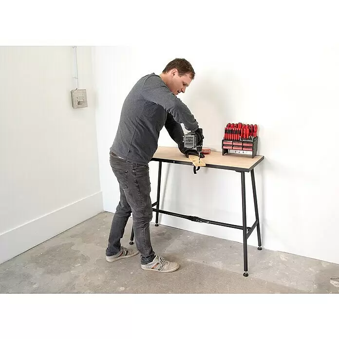 Rikta Werkbank klappbar - L x B: 100,5 x 50,6 cm, Arbeitshöhe: 84 cm, Belastbarkeit: 150 kg, Schwarz, Holz - Mann bohrt Holz auf stabiler schwarzer Werkbank mit Holzplatte, daneben ein umfangreiches Schraubendreher- und Bit-Set.