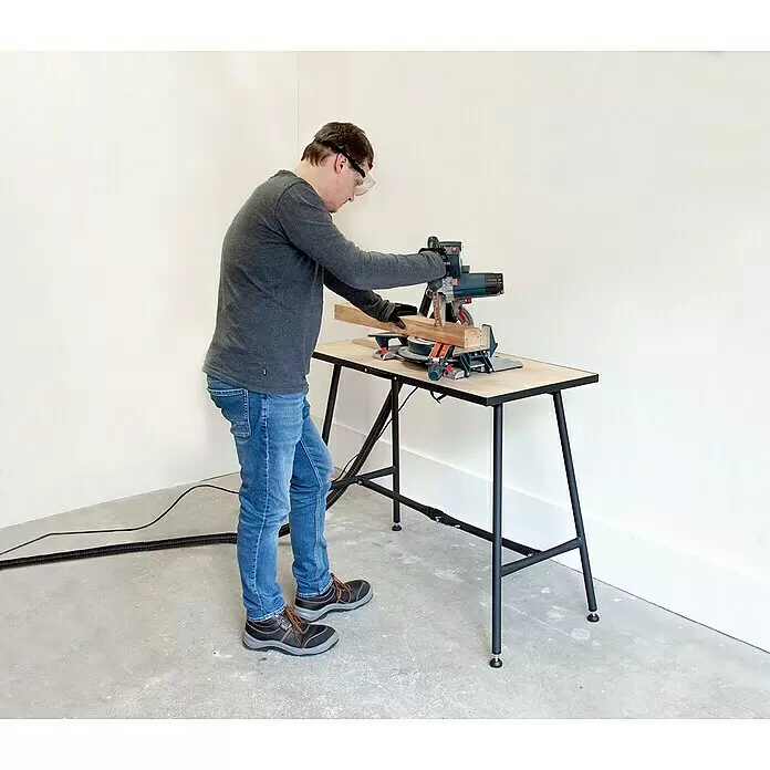 Rikta Werkbank klappbar - L x B: 100,5 x 50,6 cm, Arbeitshöhe: 84 cm, Belastbarkeit: 150 kg, Schwarz, Holz - Mann benutzt eine Gehrungssäge auf einer hellen Holzwerkbank, um Holz zu schneiden.