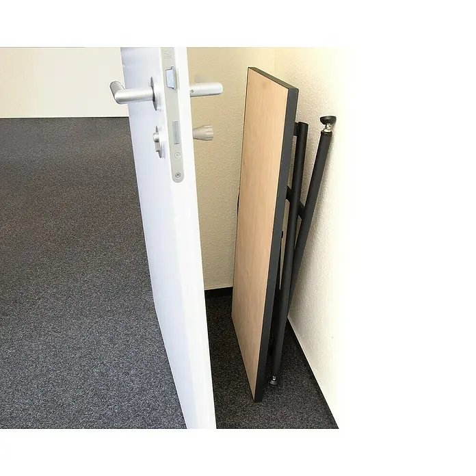 Rikta Werkbank klappbar - L x B: 100,5 x 50,6 cm, Arbeitshöhe: 84 cm, Belastbarkeit: 150 kg, Schwarz, Holz - Diese Aufnahme zeigt einen zusammengeklappten Klapptisch mit schwarzem Gestell und hellbrauner Tischplatte, der platzsparend an einer Wand steht.