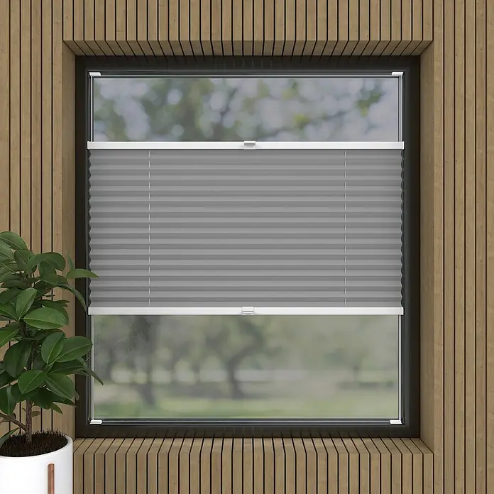Expo Ambiente Plissee Thermo - B x H: 50 x 130 cm, Grau - Graue Plissee-Jalousie mit weißem Rahmen vor einem Fenster mit Blick auf eine grüne Landschaft, umgeben von einem Holzrahmen.