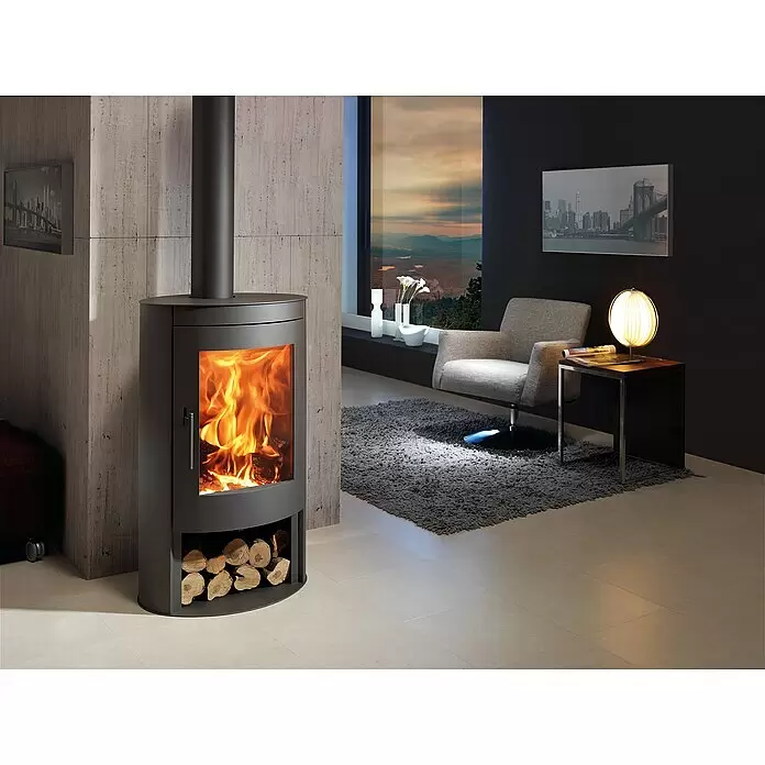 Panadero Kaminofen Oval Eco - 8,7 kW, Raumheizvermögen: 270 m³, Material Verkleidung: Stahl, Schwarz - Dunkelgrauer freistehender Kamin mit brennendem Feuer und Holzscheiten darunter, in modernem Wohnzimmer.