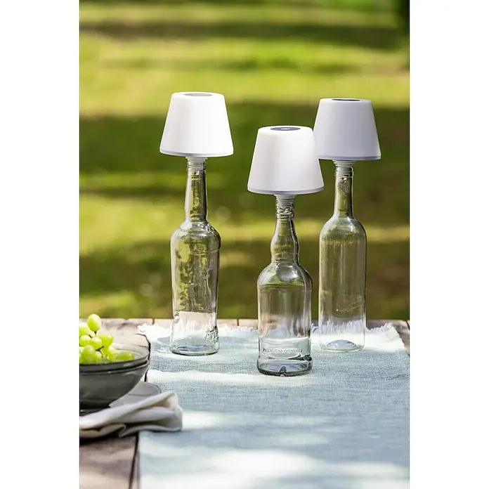 REV LED-Flaschenleuchte mit Akku Lamprusco - 2 W, Warmweiß, Ø x H: 11,5 x 13,8 cm, Weiß, Akkubetrieben - Drei Glasflaschenleuchten mit weißen Lampenschirmen auf einem Holztisch im Freien, die eine stimmungsvolle Beleuchtung für den Garten oder die Terrasse bieten.