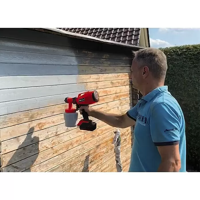 Einhell PXC 18V Sistema de pulverización de pintura de batería TC-SY 18/60 Li-Solo - 18 V, Sin batería - Hombre pinta cobertizo de madera gris claro con pistola de pintura roja.