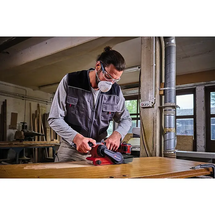 Einhell Professional PXC 18V Lijadora de banda de batería TP-BS 18/457 Li BL - Solo - 18 V, Sin batería, Velocidad de cinta: 120 m/min - 250 m/min - Un hombre lija una tabla de madera con una lijadora de banda roja Einhell, rodeado de equipo de taller.