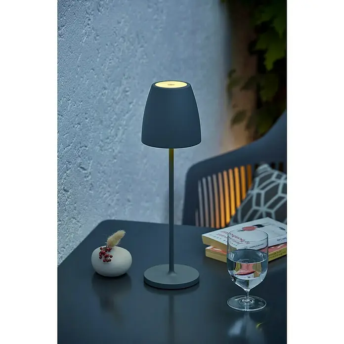 Lavida Akku-Tischleuchte Silea - 3,3 W, Anthrazit, Lichtfarbe: RGBW - Eine stilvolle, akkubetriebene Tischleuchte in Grau steht auf einem Tisch mit Dekorationen und einem Glas Wasser, ideal für gemütliche Abende.