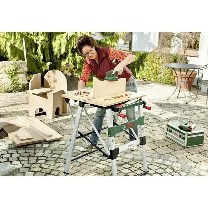 Bosch Werkbank PWB 600 - L x B: 680 x 680 mm, Arbeitshöhe: 834 mm, Belastbarkeit: 200 kg - Frau schleift Holz auf einer Bosch PWB 600 Werkbank im Freien, um ein Holzprojekt zu verfeinern.