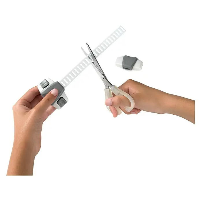Inofix Cierre para muebles 5102-2- - Blanco/gris, Tipo de montaje: Pegado - Una mano usa tijeras para cortar una correa de bloqueo de armario blanca y ajustable mientras otra mano sujeta el mecanismo de bloqueo.