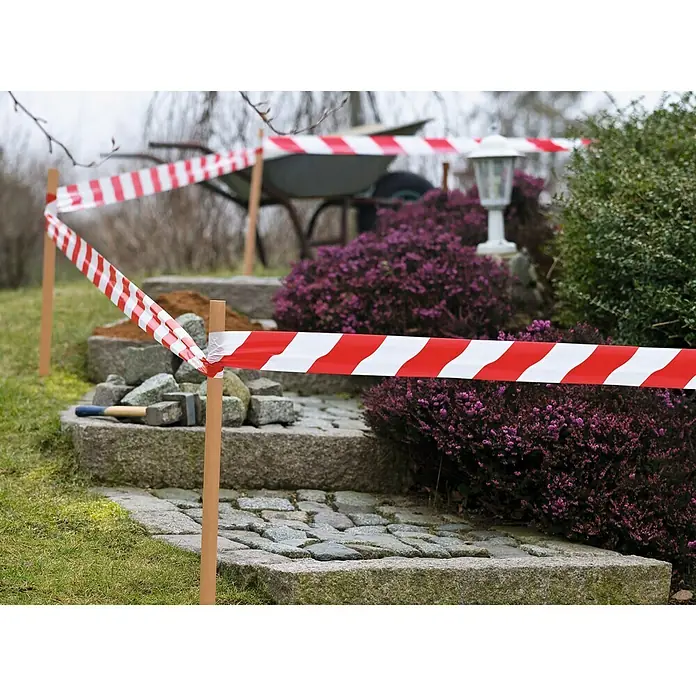Tesa Cinta de señalización Signal - 100 m x 80 mm - Cinta de construcción rodea una zona con adoquines y jardinería, con herramientas.