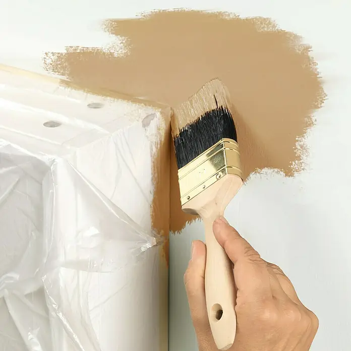 Tesa Easy Cover Plástico protector Universal - Incoloro, 25 m x 55 cm - Aplicación de pintura con brocha: Una mano pinta una pared con una brocha en un tono beige cálido, y la aplicación de la pintura es visible.