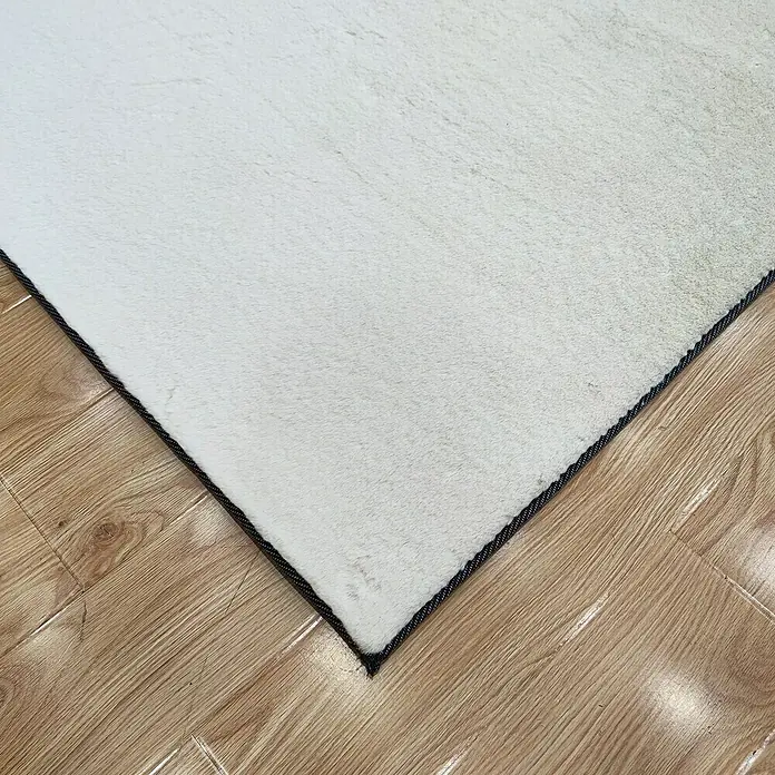 Alfombra de pelo corto Tiana - Crudo, 110 x 60 cm - Primer plano de una alfombra blanca con un borde oscuro colocada sobre un suelo de madera.