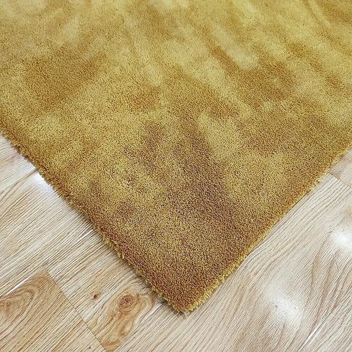 Alfombra de pelo corto Toscana - Mostaza, 230 x 160 cm, 100% Poliéster - Primer plano de una alfombra de color dorado con una textura suave sobre un suelo de madera.