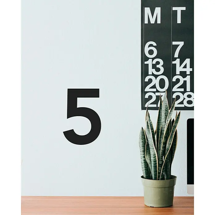 Vinilo de pared Número 5 - 4 x 9 cm, Negro, Plástico - La imagen muestra una planta en una maceta verde, con un número cinco negro en la pared y un calendario al fondo.