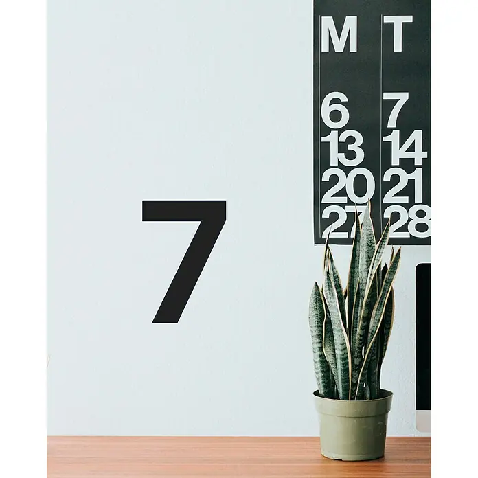Vinilo de pared Número 7 - 4 x 9 cm, Negro, Plástico - Planta de interior en una maceta verde delante de una pared con el número 7 y un calendario con fechas.