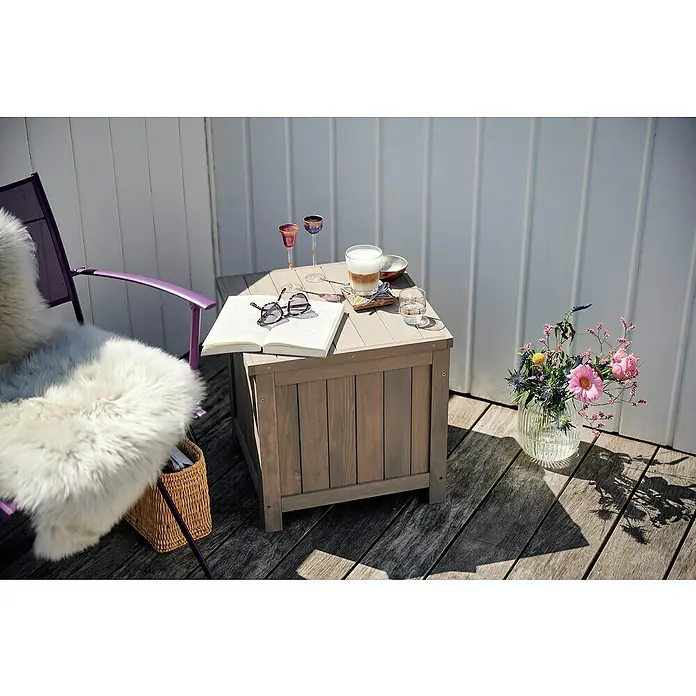 Butenkist Garten-Aufbewahrungsbox HILLE - Warm Grey, Holz - Ein stilvoller Outdoor-Bereich mit einem grauen Holztisch, dekoriert mit Getränken, Buch und Blumen, neben einem lila Stuhl mit Fell.