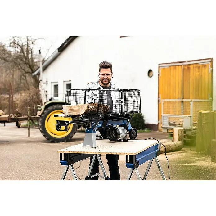 Scheppach Holzspalter LS700UG mit Untergestell - 7 t, Max. Spaltgutlänge: 52 cm - 200 mm - Ein Mann bedient einen Scheppach Holzspalter auf einem Werktisch, um Holz zu verarbeiten, mit einem Traktor im Hintergrund.