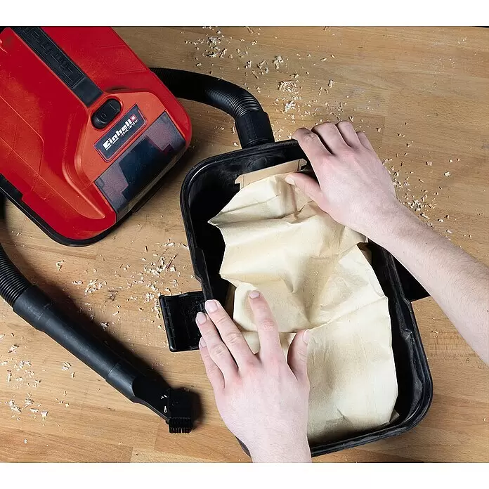 Einhell Staubbeutel - 5 Stk. - Hände setzen einen braunen Papierstaubbeutel in den schwarzen Behälter eines rot-schwarzen Einhell-Saugers ein, umgeben von Holzspänen.