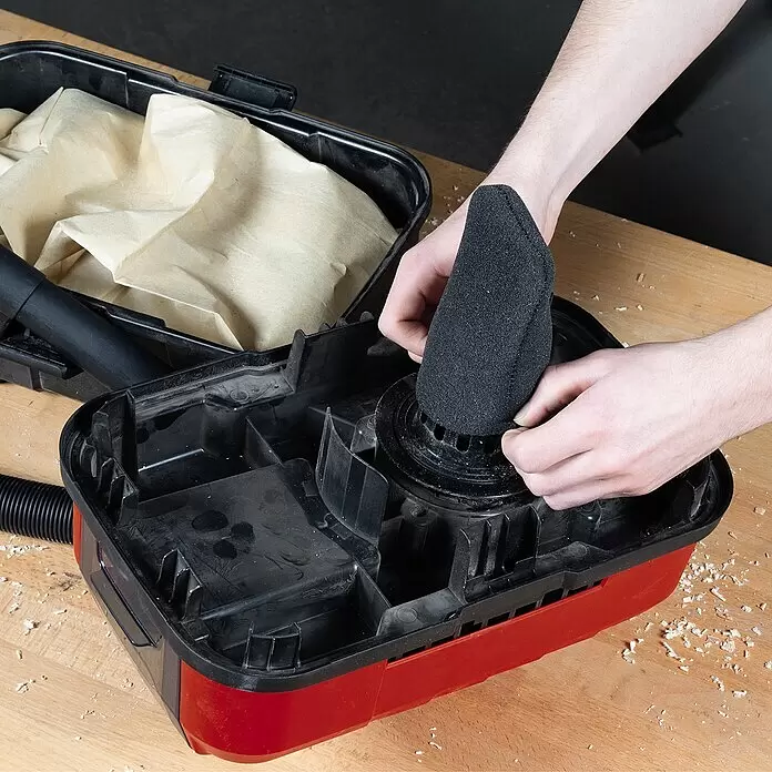 Einhell Schaumstofffilter - 5 Stk., Passend für: Einhell Nass-Trockensauger TE-VC 18/10 Li-Solo - Hände setzen einen schwarzen Schaumfilter in einen rot-schwarzen Werkstattsauger mit Papiertüte, umgeben von Holzspänen.