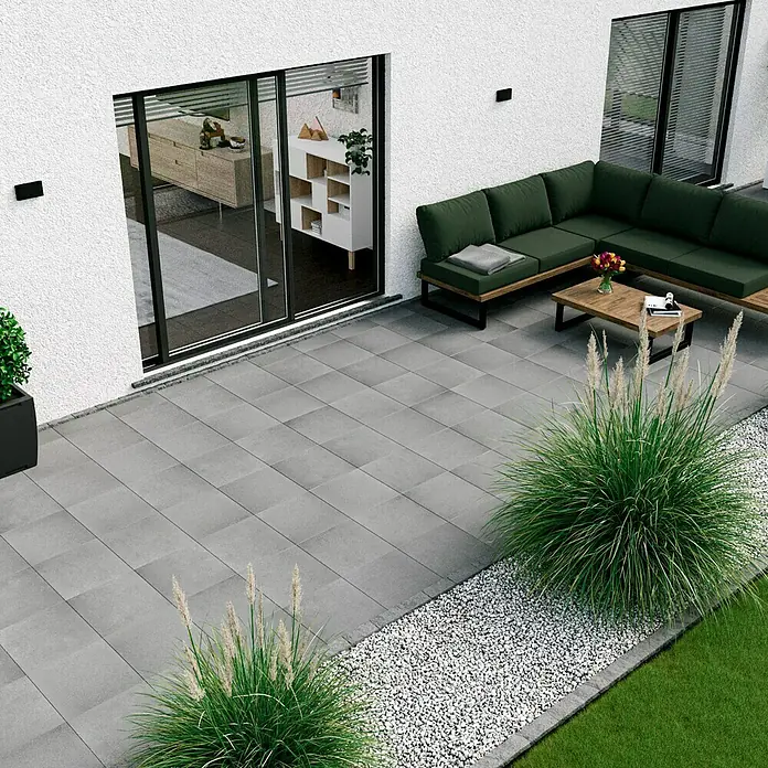 Terrassenplatte Titanio Glatt - 40 x 40 x 4 cm, Grau, Beton - Eine moderne Terrasse mit grauen Pflastersteinen, einer grünen Couch und dekorativem Ziergras, die ein stilvolles Ambiente im Freien schafft.