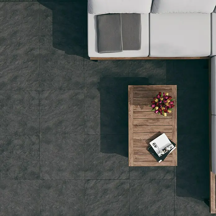 Terrassenfliese Atakama - 59,3 x 59,3 x 2 cm, Grau, Matt - Die Aufnahme von oben zeigt ein modernes Outdoor-Set mit einem grauen Fliesenboden, einem Couchtisch aus Holz und einem weißen Sofa mit Kissen.