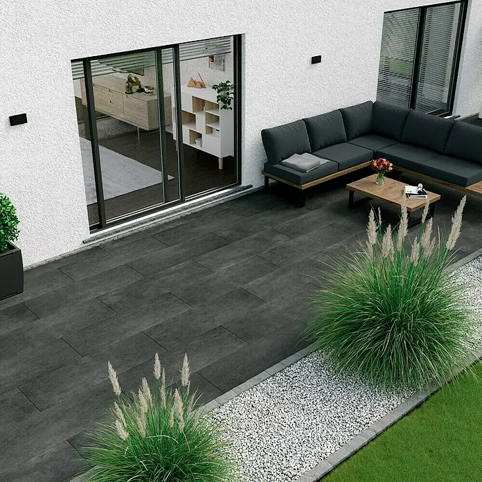 Esterno 20 T by Palazzo Terrassenfliese Basalt - 40 x 120 x 2 cm, Schwarz, Matt - Bild einer Außenterrasse mit anthrazitfarbenen Steinplatten, einer grauen Lounge-Ecke und dekorativen Ziergräsern für ein modernes Ambiente.