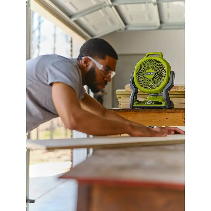Ryobi ONE+ Akku-Ventilator RF18-0 - 18 V, Ohne Akku - Ein Handwerker arbeitet an einem Tisch mit einem grünen Ryobi-Ventilator im Hintergrund, der für Kühlung sorgt.
