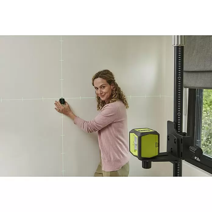 Ryobi Kreuzlinienlaser RBCLLG2 mit Skalamarkierung - Max. Arbeitsbereich: 15 m - Eine Frau nutzt ein grün-schwarzes Laser-Nivelliergerät, das grüne Kreuzlinien zur Ausrichtung an eine Wand projiziert.