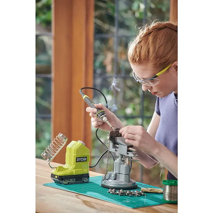 Ryobi ONE+ Akku-Lötkolben RSI18-0 - 18 V, Ohne Akku - Nahaufnahme einer Person, die mit einem Ryobi-Lötkolben an einem kleinen Projekt arbeitet, wobei der Lötkolben und die dazugehörige Basis im Fokus stehen.