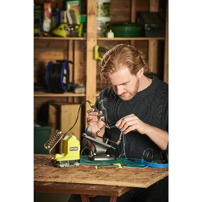 Ryobi ONE+ Akku-Lötkolben RSI18-0 - 18 V, Ohne Akku - Ein Mann arbeitet mit einem RYOBI Lötkolben an einem Tisch in einer Werkstatt, während Rauch aufsteigt. Werkzeuge und Zubehör sind auf dem Tisch verteilt.