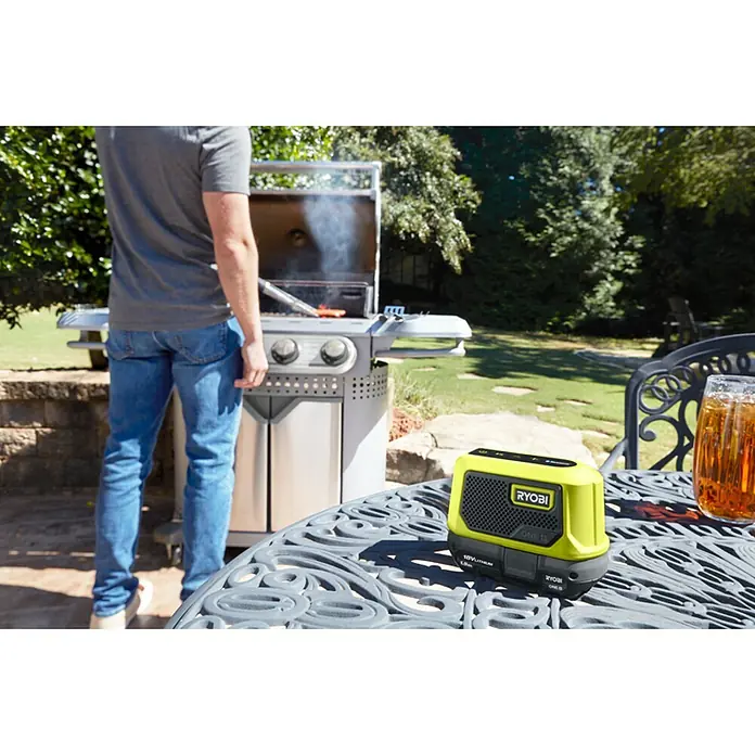 Ryobi ONE+ Akku-Bluetooth-Radio RBTM18-0 - 5 W, Ohne Akku - Der gelbe Ryobi ONE+ Lautsprecher steht auf einem Tisch während jemand grillt.