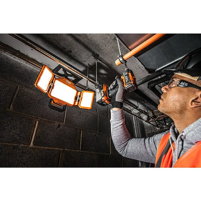 AEG Pro 18V LED-Arbeitsleuchte BLP18 - 18 V, 3.000 lm, Ohne Akku - Ein Installateur befestigt mit einem AEG-Akkuschrauber eine Rohrschelle an einer Decke, beleuchtet von einer AEG-Arbeitsleuchte.