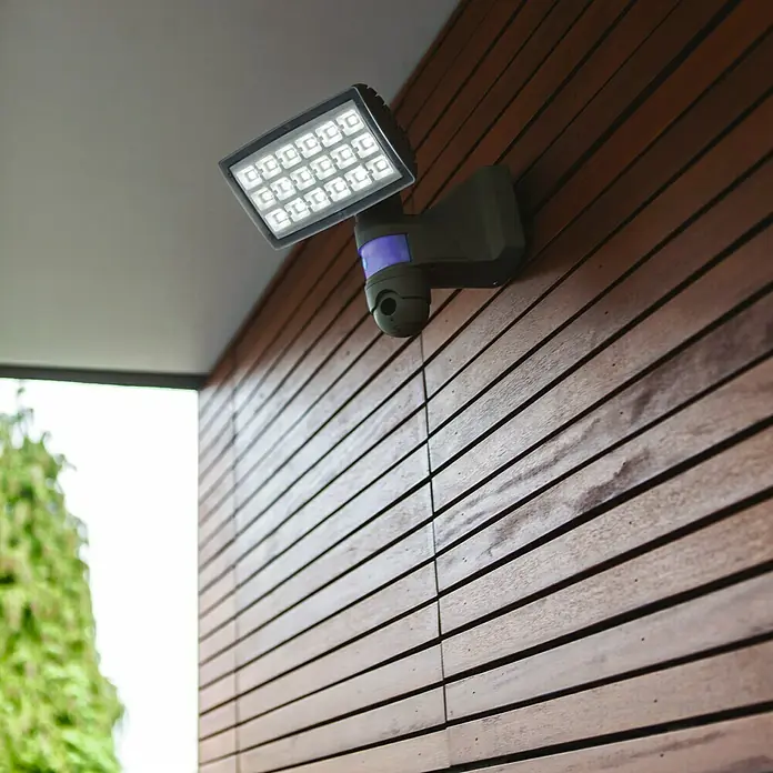 Lutec Proyector LED Peri con sensor y cámara - 25 W, L x An x Al: 22,6 x 20 x 27,1 cm, Gris, Blanco frío - Primer plano de una moderna luz LED exterior con sensor de movimiento montada en una pared de madera, que garantiza seguridad e iluminación elegante.