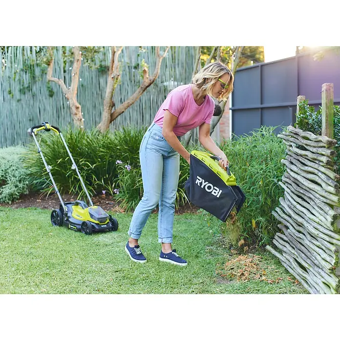 Ryobi ONE+ Cortacésped de batería RLM18X33B40 - 18 V, 1 batería, 4 Ah, Ancho de corte: 33 mm - Una mujer está vaciando una bolsa de recogida de hierba de un cortacésped inalámbrico RYOBI en un jardín bien cuidado, con el cortacésped al fondo.