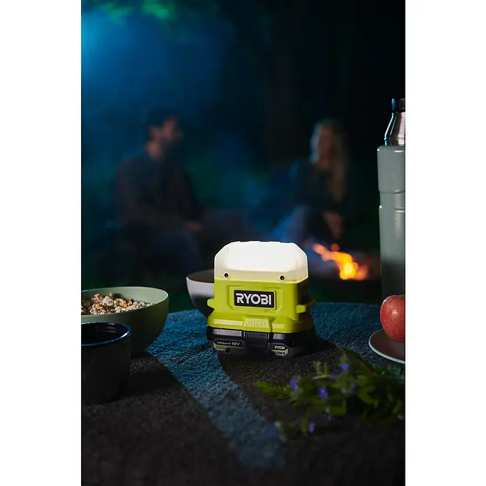 Ryobi ONE+ LED-Leuchte RLC18-0500 lm, Ohne Akku - Ryobi ONE+ Akku-Laterne beleuchtet einen Tisch bei einem Campingausflug, während Personen sich am Lagerfeuer entspannen.