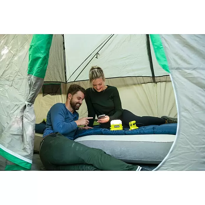 Ryobi ONE+ LED-Leuchte RLC18-0 - 500 lm, Ohne Akku - Ryobi gelbe Laterne und Wechselrichter beleuchten ein Zelt, während Personen Geräte auf einer Luftmatratze aufladen.